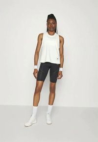 Top sportiv fără mâneci de culoare gri deschis, şorturi negre strâmte cu model perforat, brățări albe pentru încheietură, pantofi albi, fundal neutru.