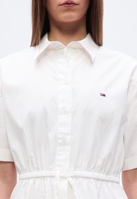 Γυναίκα με λευκό φόρεμα πουκάμισο με κοντά μανίκια και ελαστική μέση, που φέρει ένα μικρό κόκκινο, λευκό και μπλε λογότυπο στο στήθος.