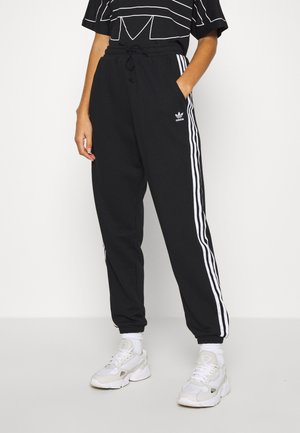 Persoon die zwarte Adidas trainingsbroek draagt met witte strepen langs de zijkanten, witte sneakers en een zwart met patroon bedrukte shirt met korte mouwen.