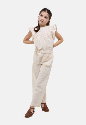 Jeune fille portant un haut sans manches crème à volants et un pantalon assorti avec de grandes poches à l'avant, debout les mains derrière le dos et regardant vers le haut.