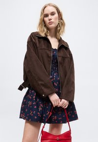 Ξανθιά γυναίκα που φοράει ένα καφέ φαρδύ μπουφάν πάνω από ένα σκούρο μπλε φόρεμα με κόκκινα λουλούδια, κρατώντας μια κόκκινη τσάντα με τα δύο χέρια.