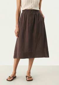 Jupe en lin marron avec une taille élastique, un détail de bouton et des poches latérales. Tombe au-dessous du genou avec une coupe décontractée. Portée avec des tongs.