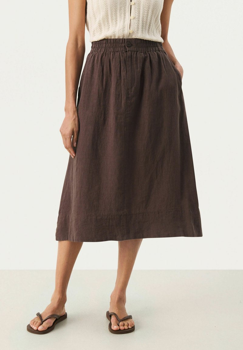 Jupe en lin marron avec une taille élastique, un détail de bouton et des poches latérales. Tombe au-dessous du genou avec une coupe décontractée. Portée avec des tongs.