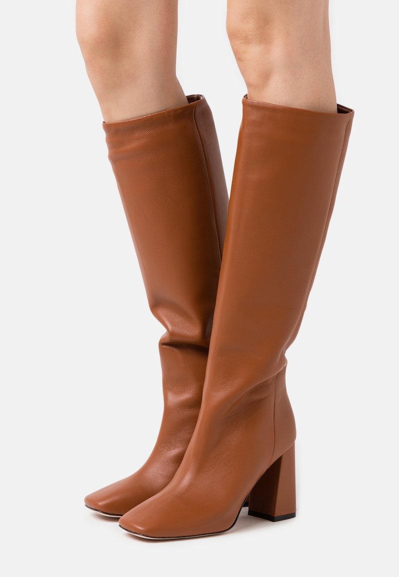 Bottes en cuir marron, hautes jusqu'au genou, avec un bout carré, un talon bloc et une texture lisse. Elles présentent un design ajusté sans matériel visible.
