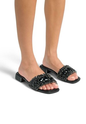 Black slide sandals featuring a wide, embellished strap with large black gems and smaller beads, set on a low block heel.