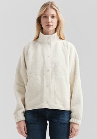 Veste en polaire crème avec un col montant, une fermeture à boutons et deux poches avant. Présente une surface douce et texturée ainsi qu'une coupe décontractée.