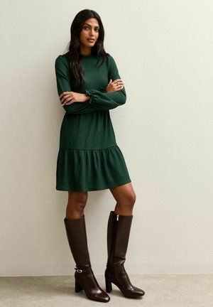 Green, ribbed long-sleeve dress with a tiered skirt; paired with knee-high, brown leather boots featuring block heels and silver hardware accents.