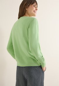 Femme souriante, portant un pull léger à manches longues vert clair et un pantalon gris foncé, debout les mains dans les poches devant un fond uni.