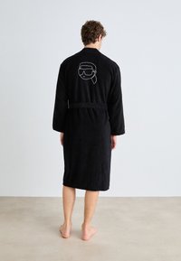 Robe de banho em tecido felpudo preto, com mangas longas, cinto na cintura e uma estampa nas costas de um rosto usando óculos escuros. Textura suave, design até aos joelhos.