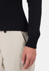 Schwarzer gerippter Pullover mit tailliertem Design, versehen mit einem strukturierten Strick und engen Bündchen, kombiniert mit hellbeigen Hosen.