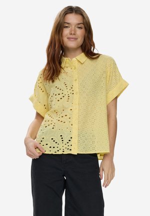Jeune femme portant une chemise jaune à œillets à manches courtes et amples, boutonnée, et un pantalon noir, debout devant un fond blanc uni.