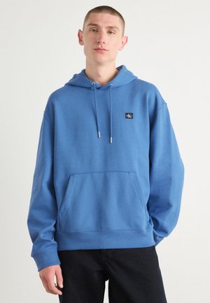 Blauwe katoen hoodie met een voorzak, een trekkoordkap en een logopatch op de borst. Ribgebreide manchetten en onderkant maken het ontwerp af.