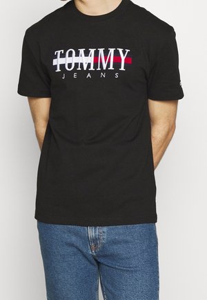Homme portant un t-shirt noir avec le logo "TOMMY JEANS" en blanc sur une bande rouge et blanche, associé à un jean denim bleu, debout sur fond blanc.