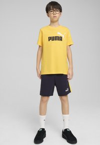 Gul bomull t-shirt med svart "PUMA"-logga, parat med marinblå shorts med en gul accent. Bärs med svarta sneakers och vita strumpor.