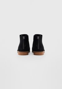 Unilă de cizme încheiate din suede negru, cu tocuri din lemn maro, văzute din spate, având fermoare verticale pe fiecare toc.