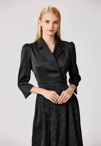 Femme blonde portant une robe blazer en satin noir avec des épaules bouffantes et des manches 3/4, se tenant devant un fond léger uni.