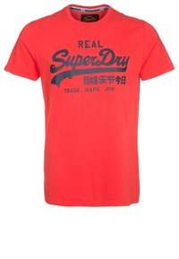 Camiseta de algodón roja con mangas cortas. Presenta un diseño de logo y texto de "SuperDry" en azul oscuro. Cuello redondo clásico. Textura suave.