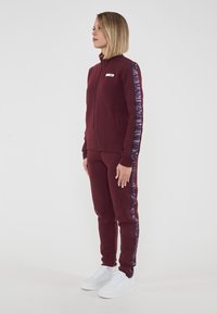 Bordowy dres wykonany z miękkiej tkaniny, składający się z kurtki na zamek i dopasowanych spodni. Wyraźne logo w purpurowym kolorze na rękawach, w połączeniu z białymi sneakersami.