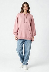 Felpa oversize rosa con coulisse, tasca frontale, orlo a coste, abbinata a jeans leggeri blu chiaro e sneakers bianche. Texture liscia.