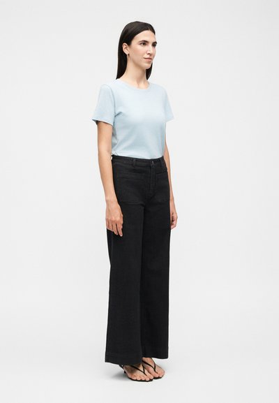 Γυναίκα που στέκεται πλάγια φοράει ανοιχτό μπλε κοντομάνικο πουκάμισο, μαύρο παντελόνι με φαρδιά μπατζάκια και ψηλή μέση, και μαύρα σανδάλια σαγιονάρα.