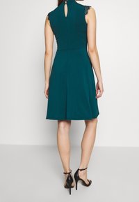 Robe turquoise avec des manches courtes en dentelle, un dos en forme de clé et une jupe évasée, complétée par des talons noirs à brides. Texture lisse.