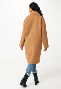 Cappotto di lana color cammello con un ampio colletto, maniche lunghe e una vestibilità rilassata, abbinato a jeans blu e stivali neri con tacco.