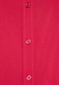 Camisa rosa de botones con una textura suave, que presenta una fila de cuatro botones, costuras visibles y un acabado ligeramente brillante.