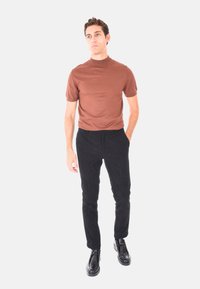 Uomo in piedi che indossa un maglione a collo alto corto marrone, pantaloni in velluto a coste neri e scarpe in pelle nere su uno sfondo bianco semplice.