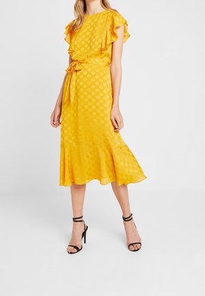 Femme portant une robe midi jaune avec des manches courtes à volants, une ceinture nouée à la taille et des sandales à talons hauts noires avec bride cheville sur fond blanc.