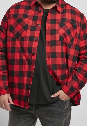 Mann trägt ein aufgeknöpftes rot-schwarz kariertes Flanellhemd über einem schwarzen T-Shirt, mit einer Hand in der Jeans-Tasche.