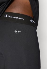 Černé sportovní šortky s hladkou texturou, které mají černý elastický pás se bílým logem "Champion" a stříbrným logem "C DRY".