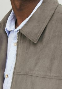 Jachetă din suede gri cu guler tăiat și închidere cu fermoar, purtată peste o cămașă în dungi albastre deschise. Prezintă un material texturat și coasere vizibile.