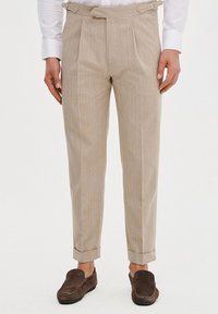 Beżowe spodnie w paski wykonane z lekkiego materiału, z plisowanym wzornictwem, mankietami oraz bocznymi klamrami. Noszone z brązowymi butami wsuwanymi.