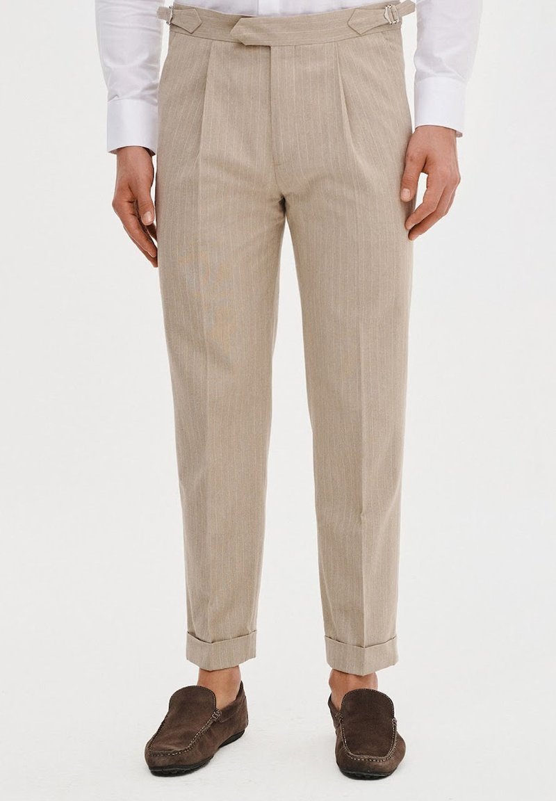 Beżowe spodnie w paski wykonane z lekkiego materiału, z plisowanym wzornictwem, mankietami oraz bocznymi klamrami. Noszone z brązowymi butami wsuwanymi.