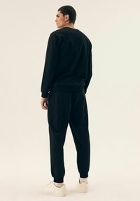 Czarna bluza i pasujące dresy wykonane z miękkiego materiału, z elastycznymi mankietami. Model stoi tyłem do kamery, ubrany w białe sneakersy.