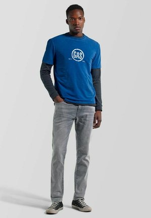 Joven de pie con una mano en el bolsillo, vistiendo camisas azules superpuestas con el logo "GAS", jeans grises y zapatillas negras sobre un fondo liso.