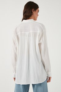 White long-sleeve shirt with a textured fabric, featuring a collar, button cuffs, and a slightly oversized fit. Notable back pleat detail.