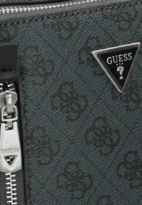 Bolso de tela texturizada negra con un diseño estampado, cremalleras plateadas y un logo en relieve triangular con "GUESS USA."