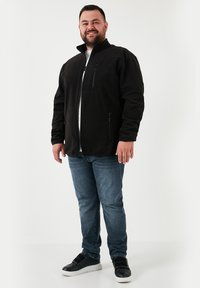 Chaqueta de forro polar negra con un bolsillo en el pecho con cremallera, cuello alto y dos bolsillos laterales, combinada con vaqueros azules y zapatillas negras.