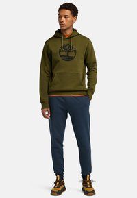 Felpa verde olivo con un logo a forma di albero nero, abbinata a pantaloni jogger blu navy. Tessuto textured e dettaglio della tasca a canguro. Calzature marroni e nere.