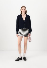 Marineblå strikket cardigan med dyb V-udskæring, parret med shorts i sort og hvidstribede mønstre. Hun holder en lille lyserød taske, iført sorte sko og sokker.