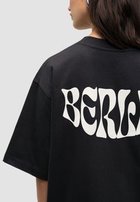 T-shirt noir surdimensionné avec un grand graphique ondulé blanc portant le mot "BERLIN" dans le dos. Fabriqué en coton doux, avec une coupe décontractée.