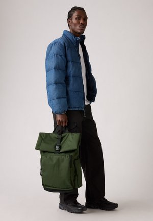 Sac à dos vert avec rabat, équipé de ferrures noires, porté à la main. Associé à une doudoune bleue et un pantalon noir, en tissu texturé.