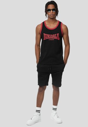 Man som bär svart och röd Lonsdale London-ärmlös tröja, svarta shorts, vita sneakers med randiga strumpor och solglasögon, står med händerna i fickorna.