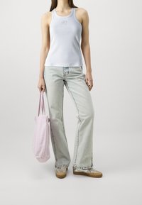 Kvinna i vit ärmlös topp, ljusblå vida jeans, beige sneakers, som håller en rosa tote-väska framför en neutral bakgrund.