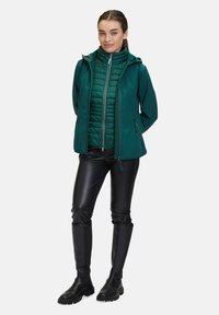 Grön vattentålig jacka med dragkedja fram och huva ovanpå en puffy grön väst; ihop med svarta åtsittande byxor och knähöga svarta stövlar.