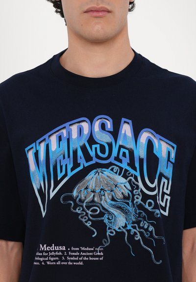 Tumesinine puuvillane T-särk, millel on sinine gradient "VERSACE" logo ja meduuside graafika, allosas on tekst, mis seletab selle tähendust.