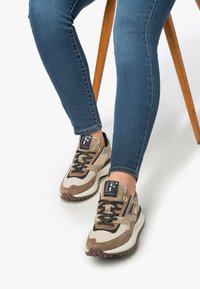 Beige and brown athletic shoes with textured fabric and smooth leather accents, featuring black laces and a logo patch on the tongue.