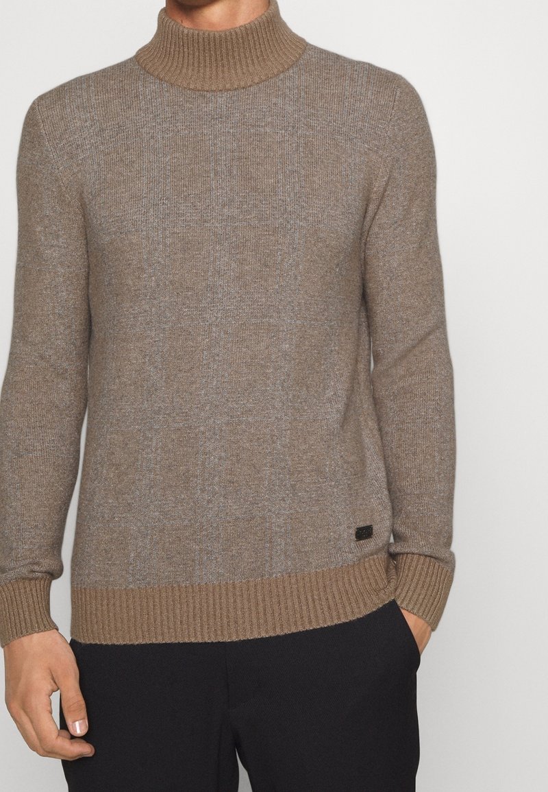 Brauner Strick-Turtleneck-Pullover mit gerippten Bündchen und Saum. Verfügt über ein dezentes Muster. Enthält ein kleines Logotag an der unteren linken Seite.