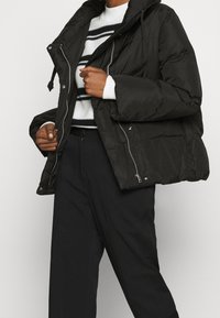 Svart puffjacka med hög krage, dragkedjestängning och fickor fram. Har en quiltad textur och lös passform, kombinerad med en randig tröja.